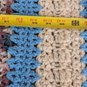 May include: Close-up of a crocheted blanket with a geometric pattern in shades of blue, cream, and mauve. The blanket is approximately 37 inches wide. A yellow measuring tape is laid across the blanket.