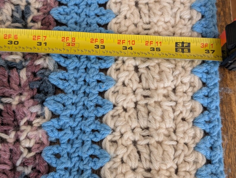 May include: Close-up of a crocheted blanket with a geometric pattern in shades of blue, cream, and mauve. The blanket is approximately 37 inches wide. A yellow measuring tape is laid across the blanket.