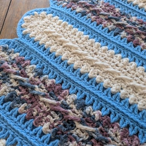 May include: A handmade crochet blanket with a scalloped edge. The blanket features a repeating pattern of light blue, cream, and muted purple sections. The crochet stitches create a textured, cozy appearance, perfect for home decor.