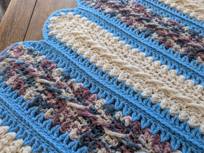 May include: A handmade crochet blanket with a scalloped edge. The blanket features a repeating pattern of light blue, cream, and muted purple sections. The crochet stitches create a textured, cozy appearance, perfect for home decor.