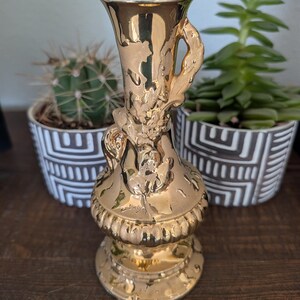 Könnte beinhalten: Eine metallisch goldene Keramikvase mit ausgestelltem Rand, einem schlanken Hals mit dekorativem Henkel und einem bauchigen Fuß mit vertikalen Rillen. Die Oberfläche hat ein reflektierendes Hochglanz-Finish mit detaillierten Reliefs.