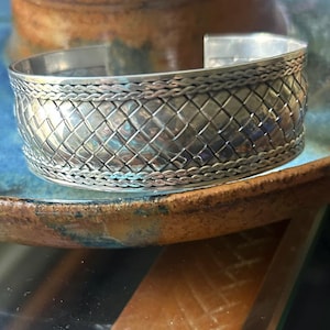 May include: A wide silver cuff bracelet featuring a central woven basket-weave pattern. The band is bordered on the top and bottom edges by a thin, braided silver wire design. The metal has a polished, reflective finish throughout the piece.