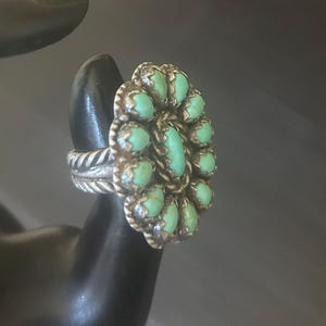 May include: A silver ring featuring a central oval turquoise stone surrounded by a cluster of ten smaller oval turquoise stones. The setting includes a twisted rope metal border and a textured, braided silver band design on the sides.