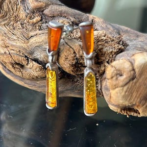 May include: A pair of drop earrings featuring two rectangular amber stones set in silver-toned metal. Each earring consists of a smaller top amber piece and a larger bottom amber piece connected by a hinge, showcasing warm orange and yellow hues.