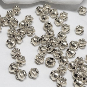 May include: A large quantity of silver metal beads with a decorative design. The beads are scattered on a white surface.
