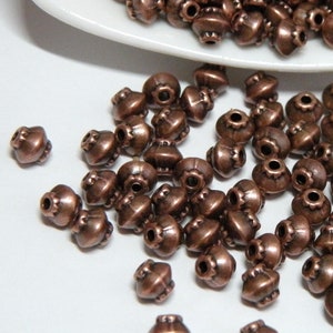 May include: A pile of copper-colored metal beads with a decorative design. The beads are round and have a small hole in the center. They are scattered on a white surface.