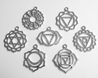 May include: Seven silver-colored chakra pendants with various geometric designs, including triangles, circles, and floral patterns. Each pendant has a small loop at the top for attaching to a chain or cord. The pendants are displayed on a white background.