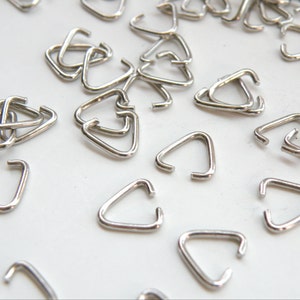 May include: A large quantity of silver-toned metal triangle jump rings. The jump rings are scattered on a white surface.