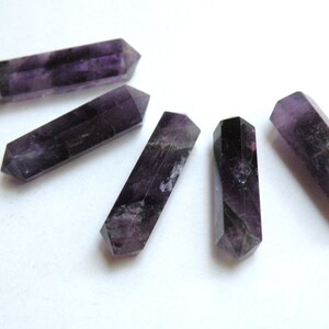 May include: Five purple amethyst crystal points, each with a double-pointed end, arranged in a semi-circle on a white surface.