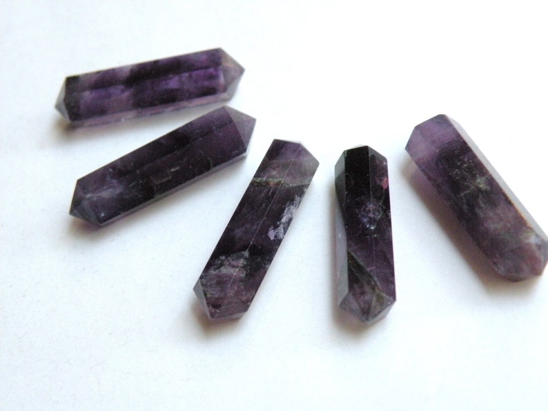 May include: Five purple amethyst crystal points, each with a double-pointed end, arranged in a semi-circle on a white surface.