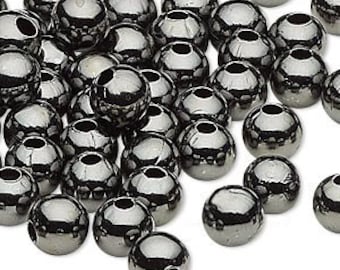 May include: A collection of shiny, silver-colored metal beads. Each bead is spherical with a small hole running through the center. The beads are scattered across a white surface, reflecting light.