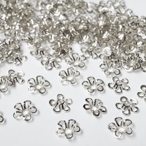 May include: A collection of small, silver-colored flower-shaped metal bead caps. The caps have a delicate, openwork design with multiple petals and a central hole. They are scattered across a white surface, suggesting their use in jewelry making or crafting.