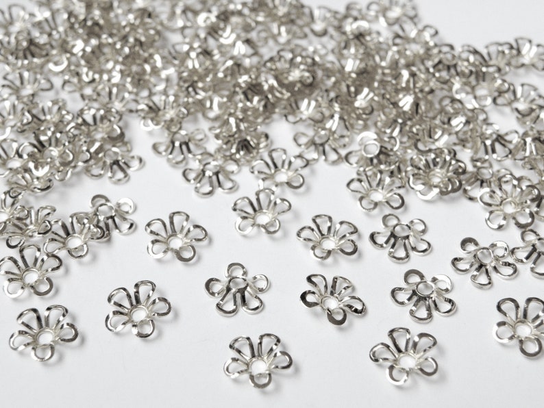 May include: A collection of small, silver-colored flower-shaped metal bead caps. The caps have a delicate, openwork design with multiple petals and a central hole. They are scattered across a white surface, suggesting their use in jewelry making or crafting.