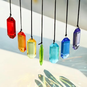 May include: A collection of colorful, faceted crystal pendants suspended from black cords. The crystals are in a rainbow of colors, including red, orange, yellow, green, blue, and purple. The crystals are hanging in front of a white background.
