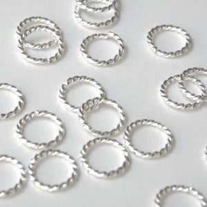 May include: A collection of silver jump rings with a twisted rope design. The rings are various sizes and are scattered on a white surface.