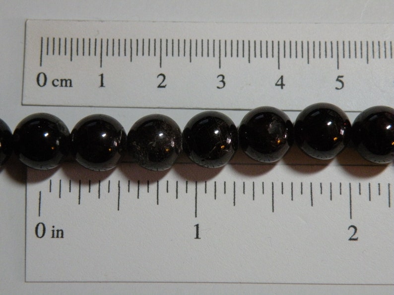 May include: A strand of black gemstone beads, each bead is approximately 1.2 cm in diameter. The beads are lined up next to a ruler, showing the size of the beads.
