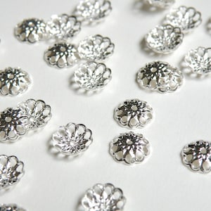 May include: A collection of silver-toned metal bead caps with a decorative, floral design. The caps are scattered on a white surface.