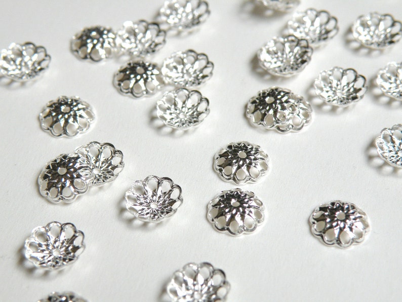 May include: A collection of silver-toned metal bead caps with a decorative, floral design. The caps are scattered on a white surface.
