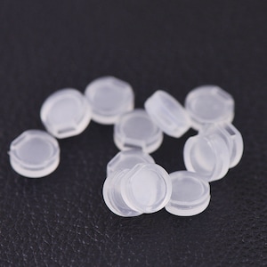 May include: A group of small, clear plastic caps. The caps are round and have a slightly raised edge. They are scattered on a black surface.