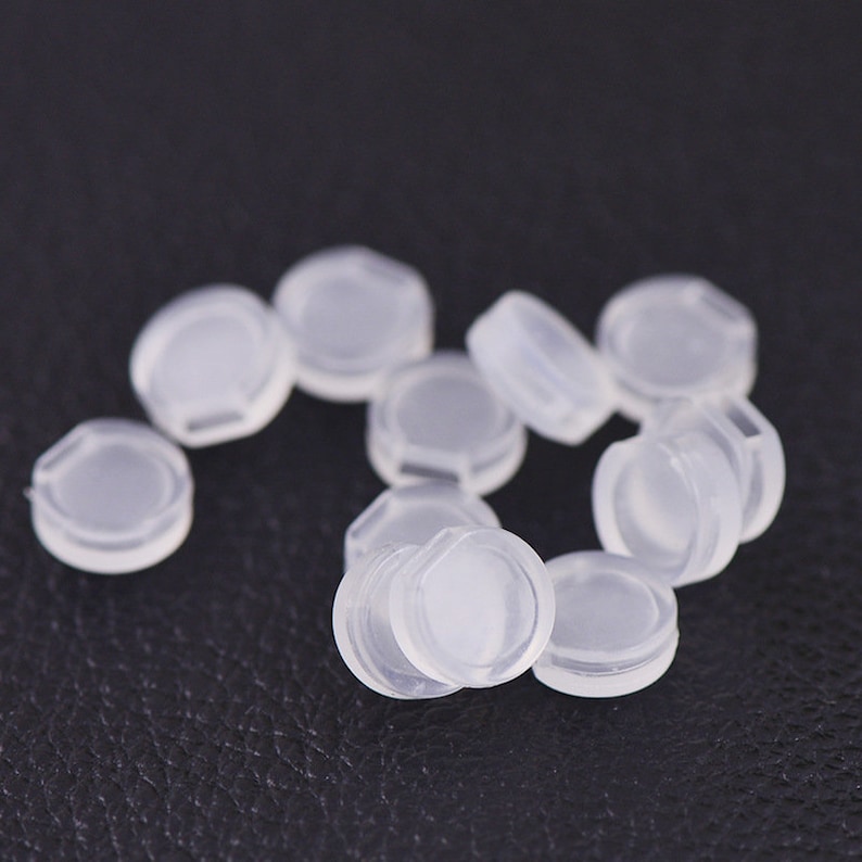 Pu&ograve; includere: Un gruppo di piccoli tappi di plastica trasparente. I tappi sono rotondi e hanno un bordo leggermente rialzato. Sono sparsi su una superficie nera.