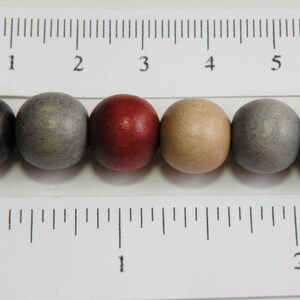 May include: A close-up of a selection of wooden beads in various colors, including black, gray, red, and tan. The beads are round and have a smooth finish. A ruler with inch measurements is in the foreground.