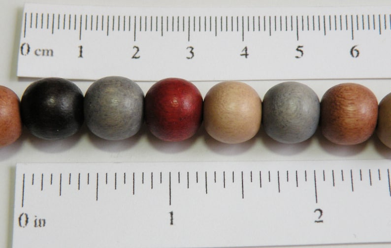 May include: A close-up of a selection of wooden beads in various colors, including black, gray, red, and tan. The beads are round and have a smooth finish. A ruler with inch measurements is in the foreground.