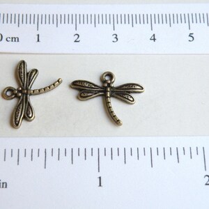 May include: Two antique gold-toned dragonfly charms, each measuring approximately 1 inch in length. The charms have detailed wings and a long, slender body.