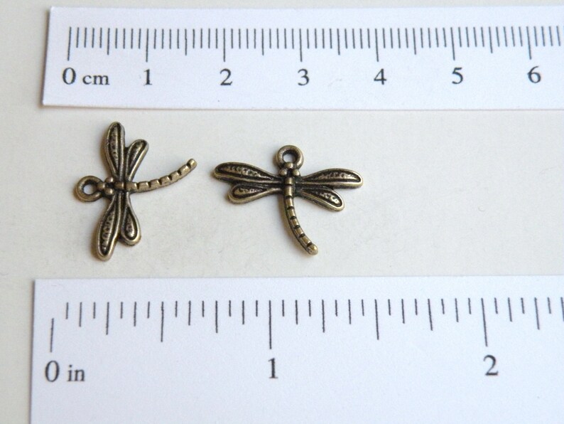 May include: Two antique gold-toned dragonfly charms, each measuring approximately 1 inch in length. The charms have detailed wings and a long, slender body.