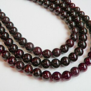 May include: A strand of dark red gemstone beads, likely garnet, strung on a cord. The beads are round and polished, and the strand is layered three times.