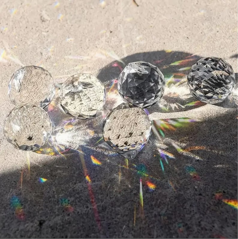 May include: Six clear crystal balls of various sizes casting rainbow reflections on a sandy surface.