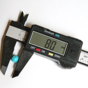 May include: A black digital caliper measuring a turquoise bead. The caliper reads 8.0 mm.