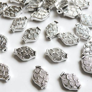May include: A collection of silver metal beads with a decorative wave pattern. The beads are oval shaped and have a hole in the center for stringing.