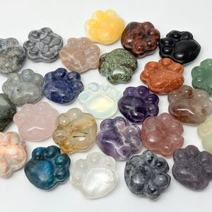 May include: A collection of polished gemstones carved into the shape of animal paw prints. The stones feature various colors and textures, including translucent white, deep blue, speckled gray, pink, purple, green, orange, and black minerals.
