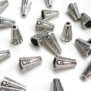 May include: A group of silver metal cone-shaped beads with an ornate design. The beads are scattered on a white surface.