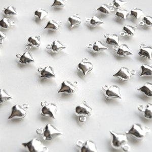 May include: A large quantity of silver heart-shaped charms with a loop at the top. The charms are scattered on a white surface.