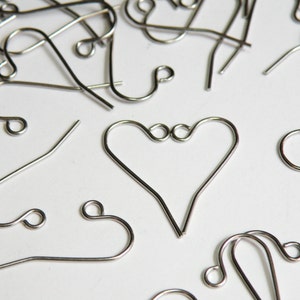 May include: A close-up of a variety of silver earring hooks, including a heart-shaped hook. The hooks are arranged on a white surface.