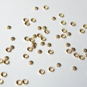 May include: Scattered on a white surface are numerous small, gold-colored, flower-shaped beads. Each bead has a central clear stone, surrounded by a scalloped edge. The beads are evenly distributed across the frame.