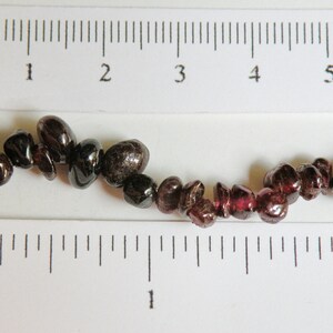 May include: A strand of dark red and black gemstone beads, measuring approximately 5 inches long, displayed on a ruler.
