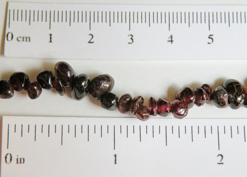 May include: A strand of dark red and black gemstone beads, measuring approximately 5 inches long, displayed on a ruler.