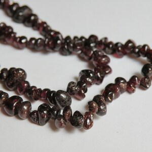 May include: A strand of dark red gemstone beads, likely garnet, strung on a clear thread. The beads are irregularly shaped and have a natural, rough texture.