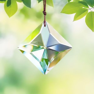 May include: A clear, faceted crystal prism, diamond-shaped, hangs from a brown cord against a blurred green background of leaves and sunlight. The crystal reflects light, creating a sparkling effect, ideal for home decor.