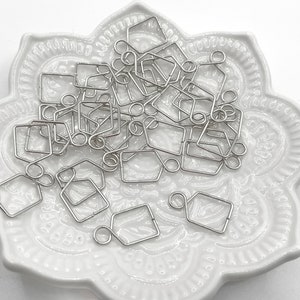 May include: A pile of silver-toned metal jump rings in a square shape with a loop on one side. The jump rings are on a white ceramic plate with a decorative design.