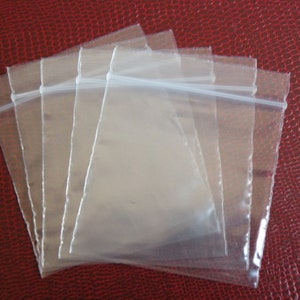May include: Four clear plastic zippered bags stacked on top of each other on a red textured surface.