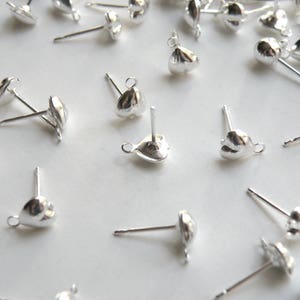 May include: A close-up of silver earring findings with a heart-shaped loop. The findings are scattered on a white surface.
