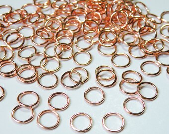 May include: A large quantity of small, rose gold jump rings. The rings are scattered on a white surface.