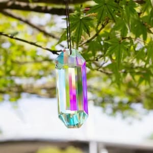 May include: A prism-shaped crystal suncatcher hanging from a thin cord. The crystal reflects a spectrum of colors, including pink, purple, blue, and green. The background features green leaves and tree branches, creating a natural setting.