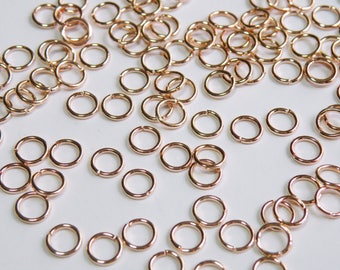 May include: A large quantity of small, rose gold jump rings. The rings are all the same size and are scattered on a white surface.
