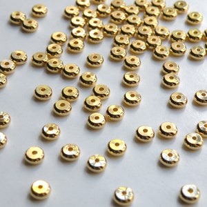 May include: A large group of gold-colored metal beads, scattered on a white surface. The beads are round with a raised edge, giving them a slightly domed appearance.