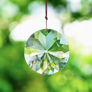 May include: A round, faceted crystal suncatcher hangs from a brown string against a blurred green background. The crystal is clear with multiple facets that reflect light, creating a sparkling effect. The suncatcher is a decorative item.
