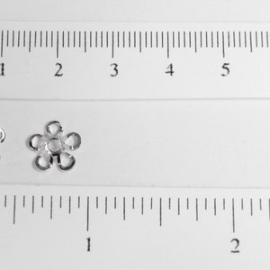 May include: Two small, silver-colored flower-shaped beads. The beads are next to a ruler showing inches and centimeters. Each bead has five petals and a central hole. The beads are likely used for jewelry making or crafting.